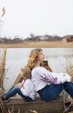 little boy and a young beautiful woman in embroidered national clothes are sitting back to back on the shore of the lake. Family, refugees, unity, support, sadness. Ukrainians are against the warの写真素材