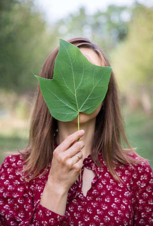Young woman holding a green leaf in her hand in front of face, hiding. faceless anonymous psychological concept. Loneliness, imagination. Place for textの写真素材
