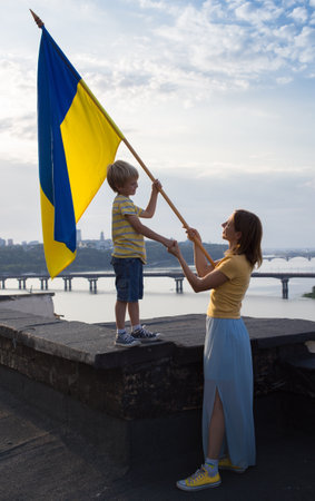 woman and child with Ukrainian flag stand on roof of house against backdrop of sky, Dnieper River in Kyiv. Patriotism, drawing attention to war in Ukraine. Support Ukraine, Hope and faith in victoryの写真素材