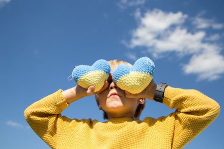 boy in a bright yellow sweater holds two knitted plush yellow-blue hearts in front of his eyes...