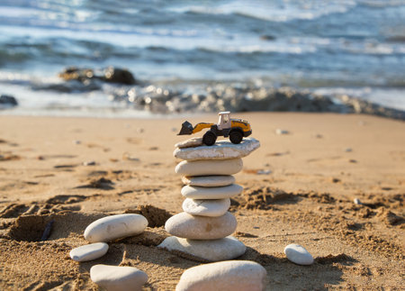 orange mini toy backhoe loader stands on tower of flat stones against backdrop of the sea. Vacation in a construction company. The concept of stability and prosperity in the construction businessの写真素材