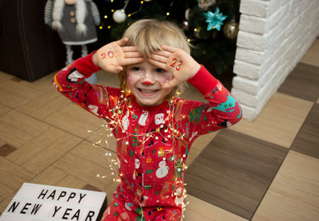 smiling joyful child in red christmas pajamas sits in front of a Christmas tree at home, numbers of...