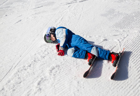 tired child in a helmet, glasses, skis and winter overalls lies on the snow. Fell into the snow, paused to rest. winter fun. Little boy learns to ski during winter holidays on a sunny cold dayの写真素材
