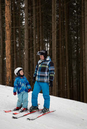 little boy learns to ski with his father or instructor stand on a snow-covered mountain slope against the backdrop of fir trees. An interesting active holiday with the family. healthy lifestyleの写真素材