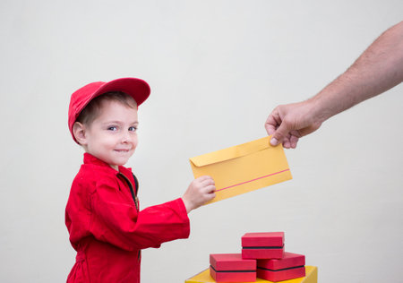 toddler boy in red overalls and cap passes yellow postal envelope to hand of adult. Express transportation, delivery of parcels and letters. postman's little helper, delivery business advertisementの写真素材