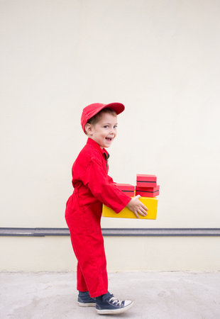 cheerful positive boy 3-4 years old in a red uniform and cap with a cardboard box in his hands. express delivery of happiness. positive feedback. space for advertising text postal businessの写真素材