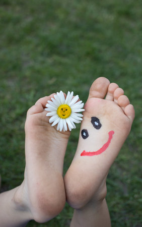 funny muzzle with big smile, drawn on bare feet of child lying on the grass. Chamomile flower between toes. Cheerful childhood. positive atmosphere, gift for mom on mother's day. selective focusの写真素材