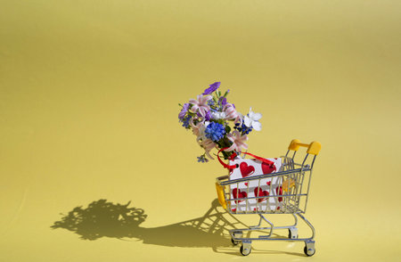 Shopping trolley, gift bag with hearts, spring flowers bouquet on yellow background. Valentine's Day, Mother's Day, Birthday. Invitations to weddings. Seasonal sales, discounts in shopping centersの写真素材