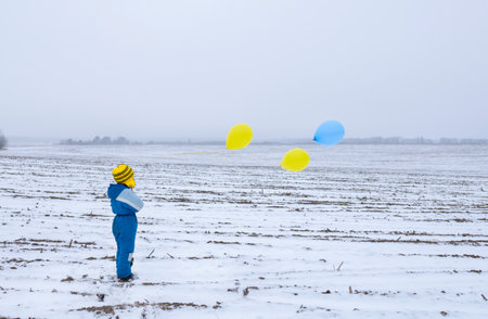 unrecognizable lonely boy standing on snowy field holding yellow blue balloons. concept of drawing attention to war, military terror in Ukraine. stolen childhood, refugee problem, nostalgia.の写真素材