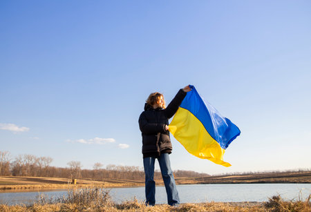 woman stands against the background of the lake and holds the Ukrainian yellow-blue flag against the sky. National symbol of freedom and independence. Support Ukraine. pride and patriotismの写真素材