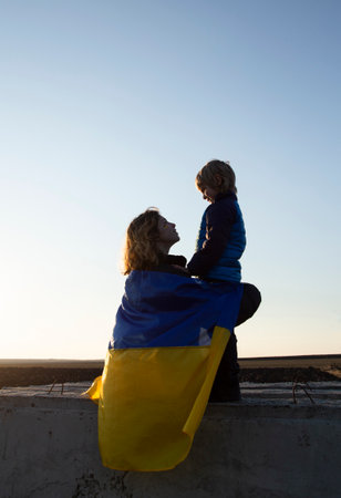 little boy and a young woman are sitting with their backs at sunset against the sky with a blue and yellow Ukrainian flag behind them. Family, refugees, unity, support, love. stand with Ukraineの写真素材