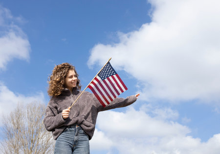 adult beautiful cheerful girl 18 years old holding an american flag in hands against blue sky on sunny day. july 4th celebration. Independence Day of the United States of America. Pride, freedomの写真素材