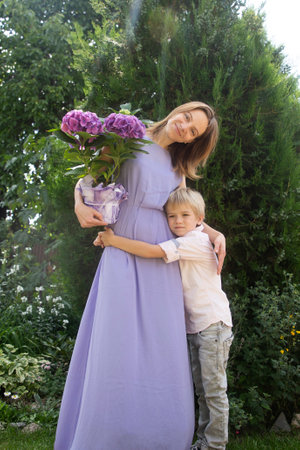 Congratulations for beloved mother on mother's day. A woman in a dress with a hydrangea in a pot and a boy hugging her stand in the garden and look at the camera. Happy family, joyful motherhoodの写真素材