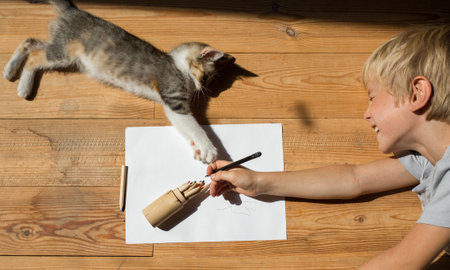 joyful child draws with pencils lying on the floor in a room on a bright sunny day in the company of a beloved kitten. Friendship and play of a child and a beloved pet. View from aboveの写真素材