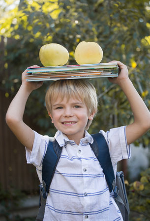 cute cheerful 6 year old boy holding stack of books and two big apples on his head, getting ready to go to school. Reading books, interesting childhood, inquisitive child, knowledge is power. book dayの写真素材