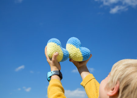 child holds in his hands two knitted plush yellow-blue hearts against a blue sky on a sunny day. the concept of patriotism, unity, support, peace for Ukraine. I am proud that I am Ukrainianの写真素材