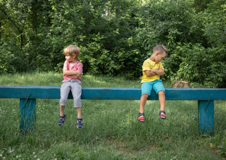 Two brothers or boys friends in casual clothes are sitting on bench offendedly turned away in different directions, sad and gloomy. Offended, dissatisfied, do not want to agree with each other.の写真素材