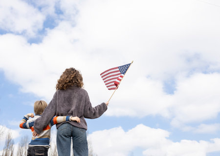 unrecognizable boy and young woman stand together with their backs holding american flag against sky. Freedom, national identity. Independence Day of the United States of America. Travel, immigrationの写真素材