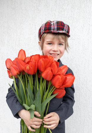 cute little boy 6 years old holds a big bouquet of red tulips on a light background. mother's day. International Women's Day. Portrait of a happy boy with a flower gift with love. Springの写真素材