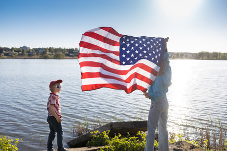 woman and child are standing on the shore of lake holding an American flag waving in hands, beautifully illuminated by the sun. patriotic holiday, Independence Day. Pride to be an American.の写真素材