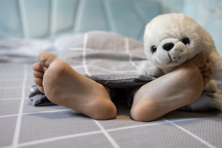 bare feet of a child on a bed under a blanket, on one foot lies a soft fluffy toy seal. Good morning. little boy sleep with pleasure with his favorite toyの写真素材