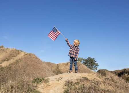 Patriotic boy 6 years old proudly raised the American flag against the blue sky. Celebration of Independence Day of the United States of America. happiness, success, freedom, national symbolの写真素材