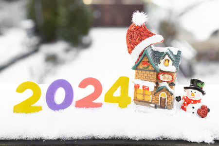 Happy New Year 2024 - numbers in snow, souvenir figurines of a snowman, a cozy cottage, a Santa hat. greeting card winter holiday mood. concept for celebrating Christmas, New Year and winter holidaysの写真素材