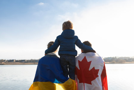 woman with Ukrainian flag, man with Canadian flag behind back and child, sitting with backs together in nature. International support and assistance to Ukraine. Problems of refugees and immigrantsの写真素材