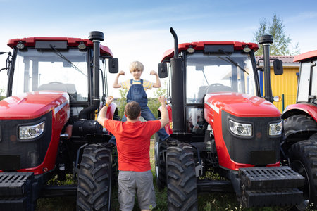 man and boy stand in front of large red new tractors at sales site. An adult and child look at agricultural machinery with interest and show its power. Father's Day, spend time together, be like dad.の写真素材