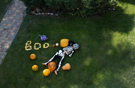 unrecognizable child in a skeleton costume lies on the grass near orange pumpkins and the words boo made from fallen leaves. preparation for the Halloween holiday. trick or treat. Top viewの写真素材