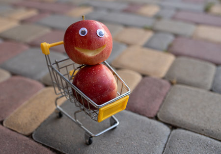 grocery cart with two red delicious apples, one with a smile cut out and eyes glued on. Funny advertisement of organic products in a supermarket. Fresh fruit shopping conceptの写真素材