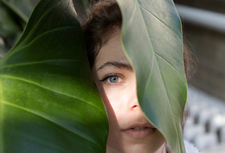 Close-up portrait of a beautiful fresh girl hidden behind large green leaves. Facial beauty. Light accent on one eye. expressive look. Tenderness, femininity, youthの写真素材