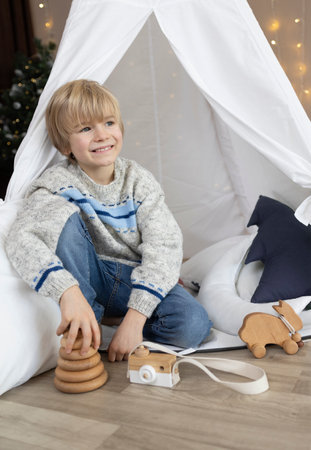 handsome smiling boy sits in a wigwam tent, with wooden toys nearby. He looks dreamily and joyfully to the side. Children's room, childhoodの写真素材