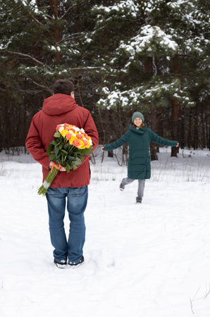 man holds a large bouquet of roses behind his back, in the background in front of him is a happy woman in love. Valentine's Day in a snowy forest or park. Romantic relationship, surprise,の写真素材