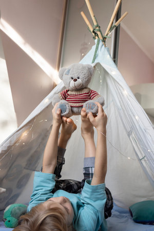 child cheerfully raising his bare feet up plays with a teddy bear while lying in a wigwam in a children's room. fantasy, interesting childhood, playing with soft toys, joyの写真素材