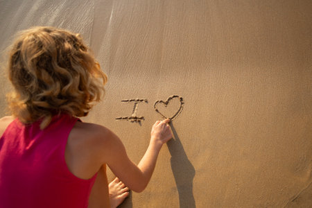 teenage girl writes on the sand I love while sitting on the sea coast. The concept of falling in love, relaxation, summer mood, vacation. travel with pleasure. digital detox. View from aboveの写真素材