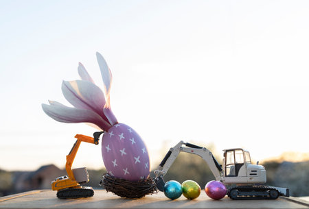 two small models of toy excavators, an eggshell with a large magnolia flower, multi-colored confetti eggs in backlight. Easter spring holiday concept, construction business greeting cardの写真素材