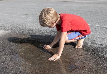 barefoot child plays with his hands in a transparent puddle on the asphalt. Sun after rain, change of weather in summer. Interesting, cheerful childhood, active lifestyleの写真素材