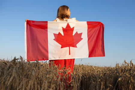 woman stands with her back among a wheat field and proudly holds a large Canadian flag, illuminated by sunlight. Canadian Independence Day. freedom, trust, pride, national symbolの写真素材