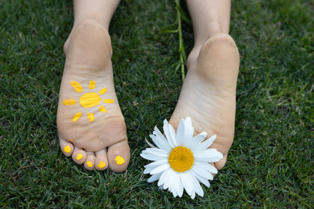 bare feet of a child lying on green grass. A daisy flower between the toes, a painted yellow sun on...