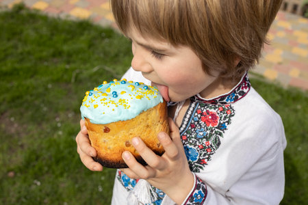 boy tastes the sweet icing from the paska. A Ukrainian child in an embroidered dress holds an Easter...