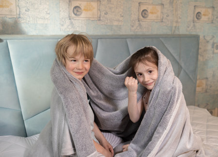 Two children are sitting next to each other on the bed. brother and sister, covered with a blanket, show their fist. reluctance to wake up or comply with a parent's request. be together.の写真素材