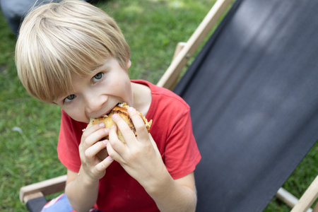 positive boy bites a sandwich during recess outdoors in the park. time for a snack on a summer walkの写真素材