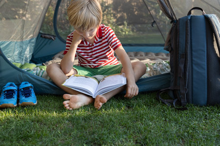 young barefoot child reads a book with interest while sitting in a tent on a warm sunny day. Summer leisure, adventure and outdoor recreation Passion for readingの写真素材