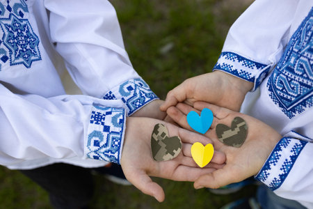 4 hearts cut from military camouflage fabric and yellow-blue paper lie on palm of two children in Ukrainian embroidered shirts. Stop war in Ukraine. With love and gratitude to soldiers of Armed Forcesの写真素材