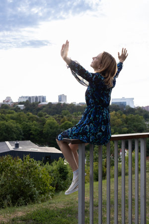 elegant woman in a dress sits on the railing with her hands raised to the sky. Fantasies, dreams,...