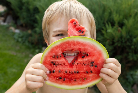 child cheerfully eats watermelon. the boy's face is hidden behind a piece of watermelon and a heart carved into it. Hello summer, good mood, delicious vitamin foodの写真素材