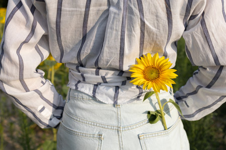 slender woman with her back in a shirt and denim shorts, a yellow sunflower in her pocket
