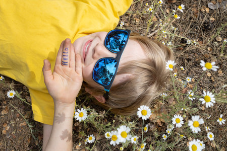 Portrait of a cute smiling boy in blue sunglasses lying among a chamomile field. Summer mood, childhood dreams, carefree joyful childhood. Hello summer, energy of nature. Earth Day. View from aboveの写真素材