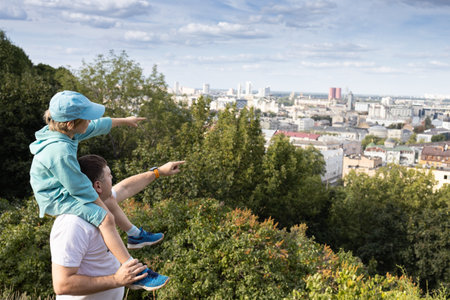 son, sitting on dad's shoulders, admires city standing on hill. Walks around the city, excursions, trips to new places. Spring, summer, outdoor recreation. happy childhood, pleasant pastime. Father's Dayの写真素材